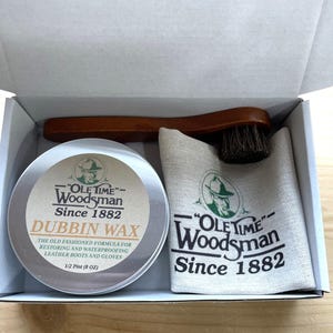Puede incluir: Un kit de cuidado del cuero en una caja, que incluye una lata de cera Dubbin, un cepillo de madera y una bolsa de lona. La lata y la bolsa muestran la marca "Ole Time Woodsman Since 1882". La cera es de 237 ml.
