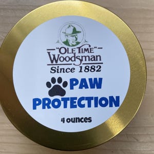 May include: A round, gold-colored tin with a white label featuring the text "Ole Time Woodsman" and "Paw Protection." The label also includes a paw print and states "4 ounces."