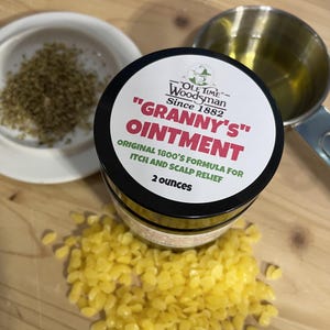 May include: A black jar of "Granny's" Ointment, with a white label and red text, sits on a wooden surface. The label reads "Original 1800's Formula for Itch and Scalp Relief" and "2 ounces". Beside it are yellow wax beads and a small white dish with brown granules.