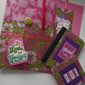 May include: A collection of stationery items with floral patterns in pink and green. Includes a pink envelope, a pen, bookmarks, and small notebooks with inspirational quotes. One tag reads "Be Kind to Yourself."