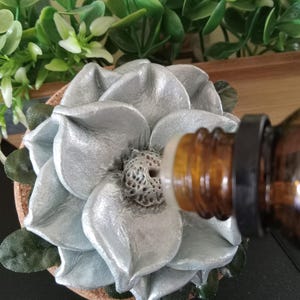 May include: A silver flower-shaped essential oil diffuser with a brown cork base. The flower has large, textured petals and a detailed center. A brown glass bottle with a black cap is positioned to dispense oil onto the diffuser.
