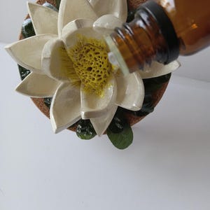 May include: A white ceramic lotus flower diffuser with a cork base and green leaves. A brown glass bottle of essential oil is being poured into the diffuser.