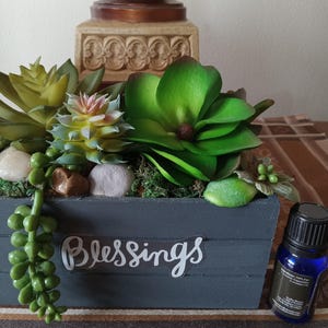 May include: A gray wooden box with the word "Blessings" on the front, filled with artificial succulents and a small blue bottle of essential oil.