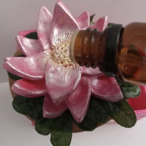 May include: A pink and white flower with green leaves and a brown essential oil bottle. The flower has multiple petals and a textured center. The bottle has a black cap.