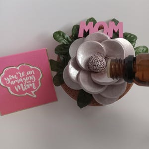 May include: A pink card with the text "You're an amazing Mom" in white lettering. A pink flower diffuser with a cork base and a wooden "MOM" sign. A brown glass bottle of essential oil is being poured into the diffuser.