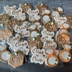 May include: Assortment of gold and white decorated cookies with various designs including rings, the words "Miss to Mrs" and the initials "C+M".