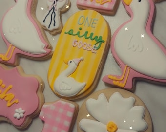 May include: Assortment of decorated sugar cookies. Designs include a goose, number one, balloons, and a flower. Colors are pink, white, yellow, and black. Some cookies have text that reads "One silly goose" and "one".