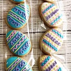 May include: A close-up of nine Easter egg-shaped biscuits decorated with colourful icing. The biscuits are arranged in two rows of five, with the bottom row slightly offset from the top row. The biscuits are decorated with a variety of patterns, including stripes, dots, and swirls. The icing is a mix of blue, purple, yellow, and pink.