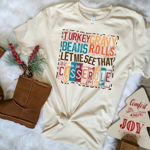 Pode incluir: Uma camiseta bege com uma estampa gr&aacute;fica de estilo vintage que diz "TURKEY GRAVY BEANS ROLLS LET ME SEE THAT CASSEROLE". O gr&aacute;fico est&aacute; em uma fonte de bloco colorida com um fundo de estampa de leopardo.
