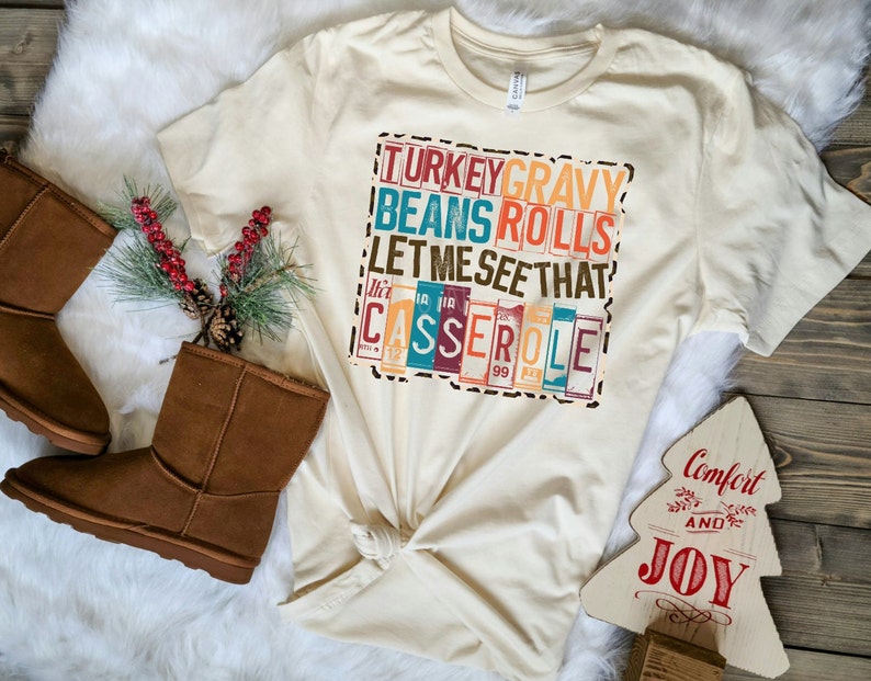 Pode incluir: Uma camiseta bege com uma estampa gr&aacute;fica de estilo vintage que diz "TURKEY GRAVY BEANS ROLLS LET ME SEE THAT CASSEROLE". O gr&aacute;fico est&aacute; em uma fonte de bloco colorida com um fundo de estampa de leopardo.