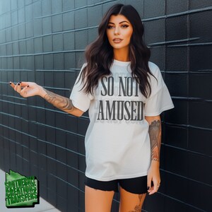 May include: A white t-shirt with the text "SO NOT AMUSED" printed in black. The shirt is being worn by a person with long dark hair and a black tattoo on their arm. The person is standing in front of a black tiled wall.