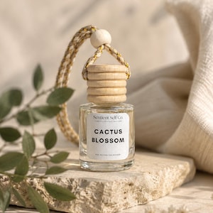Può includere: Un piccolo diffusore per auto in vetro trasparente con tappo in legno chiaro e cordino intrecciato. L'etichetta bianca riporta la scritta Sentient Self Co. e Cactus Blossom con 8 ml wooden car diffuser. Una perlina in legno è sul cordino.