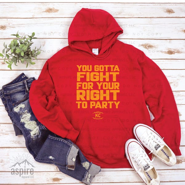 May include: A bright red hooded sweatshirt featuring the yellow text "YOU GOTTA FIGHT FOR YOUR RIGHT TO PARTY" and a small football graphic with the letters "KC" below it, paired with distressed blue denim jeans and white canvas sneakers.