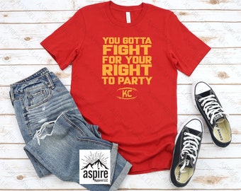 May include: Red t-shirt with the text "You Gotta Fight For Your Right To Party" in yellow with a football graphic and the letters "KC" in yellow. The shirt is laid out on a woodgrain surface with a pair of black Converse shoes and blue jeans.