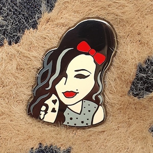 May include: A black and white enamel pin featuring a portrait of a woman with dark hair and a red bow. The woman is wearing a black and white patterned dress and has a light blue background.