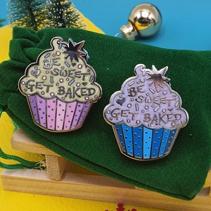 May include: Two cupcake-shaped enamel pins with the text "Be Sweet Get Baked" in a whimsical font. The pins are on a green velvet background.