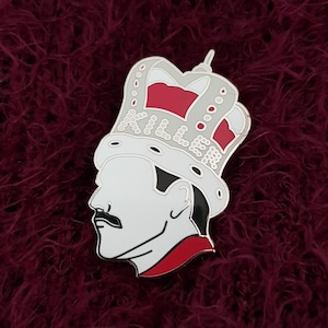 May include: A white enamel pin featuring a profile of a person wearing a red and white crown with the word "Killer" written on it.