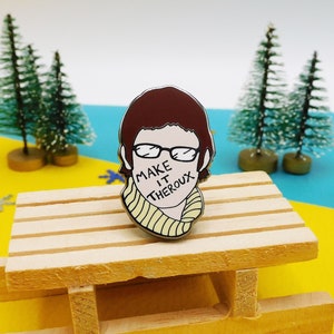 May include: A brown and white enamel pin featuring a cartoon illustration of a person with brown hair wearing glasses and a yellow scarf. The text "Make it Theroux" is written across the person's mouth.