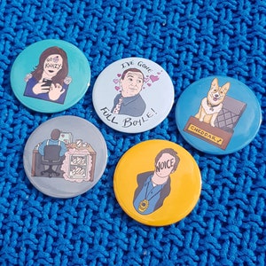 May include: Four round button badges with different designs. The first badge features a woman with a microphone and the text "Go Khazy". The second badge features a man with the text "I've Gone Full Boyle!". The third badge features a cartoon dog with the text "Cheddar". The fourth badge features a man with the text "Noice".