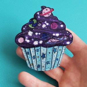 Może przedstawiać: Fioletowy i niebieski cupcake z wzorem galaktyki i białymi gwiazdami. Cupcake jest ozdobiony planetami i galaktycznym wirem.