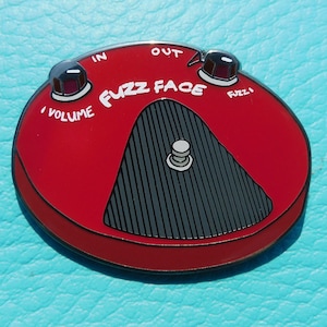 May include: Red metal pin of a Fuzz Face guitar pedal with black and white details. The pin has the words "Fuzz Face" and "Volume" on it.