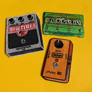 Peut inclure: Trois écussons brodés de pédales d'effets de guitare. La pédale "Big Muff" est noire, grise et rouge. Une pédale verte avec des boutons et une pédale "Phase 90" marron et orange sont également présentées.