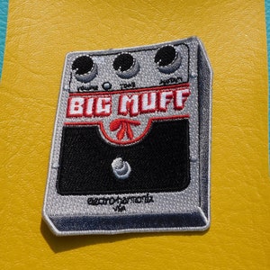 Peut inclure: Un patch brodé représentant une pédale de guitare Big Muff. Le patch est de couleur argent et noir et comporte le texte "Big Muff" en lettres rouges et blanches et le texte "Electro-Harmonix" en lettres blanches.