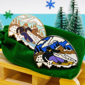 K&ouml;nnte beinhalten: Zwei Emaille-Pins mit Skifahrern vor einem schneebedeckten Bergpanorama. Die Pins sind in Gold und Silber mit blauen, wei&szlig;en und braunen Akzenten gehalten. Die Pins befinden sich auf einem gr&uuml;nen Samtbeutel.