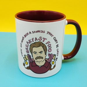 May include: White ceramic mug with a red rim and handle. The mug features a cartoon illustration of a man with a moustache and the text "There has never been a sadness that can't be cured by breakfast food."
