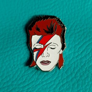 Puede incluir: Un pin de esmalte plateado con una persona de pelo rojo con un rayo pintado en la cara. Los ojos de la persona est&aacute;n cerrados y su boca est&aacute; ligeramente abierta. El fondo es azul turquesa.