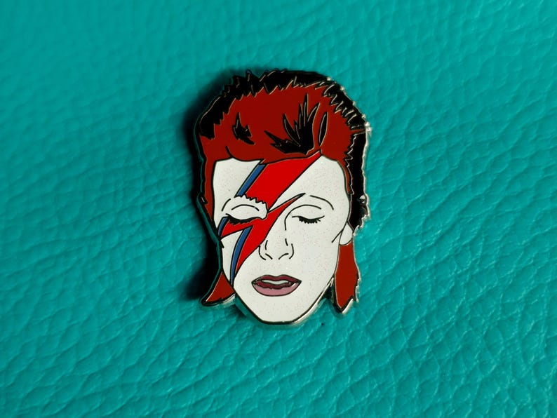 Puede incluir: Un pin de esmalte plateado con una persona de pelo rojo con un rayo pintado en la cara. Los ojos de la persona est&aacute;n cerrados y su boca est&aacute; ligeramente abierta. El fondo es azul turquesa.