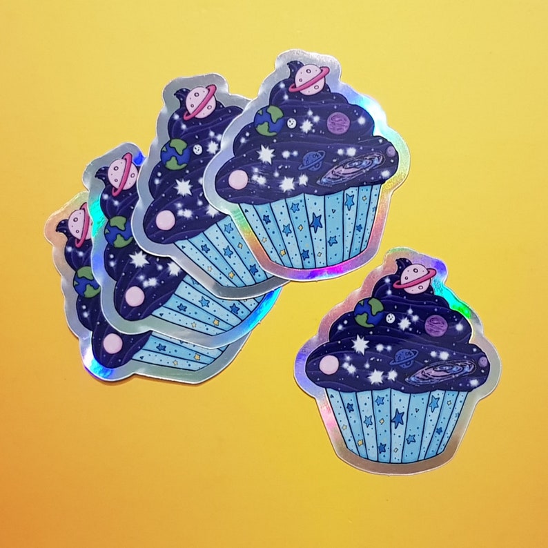 May include: Five iridescent blue and silver cupcake stickers with a space theme. Each cupcake has a dark blue frosting with white stars, planets, and a galaxy design.