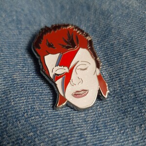 Puede incluir: Un pin de esmalte plateado con un retrato de David Bowie con su caracter&iacute;stico maquillaje de rayo. El pin es de dise&ntilde;o blanco y rojo sobre un fondo plateado.