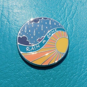 Puede incluir: Un pin redondo de color plateado con un cielo azul y nubes de lluvia en una mitad y un sol amarillo con rayos naranjas en la otra mitad. El texto "RAIN OR SHINE" est&aacute; escrito en una fuente blanca cursiva.