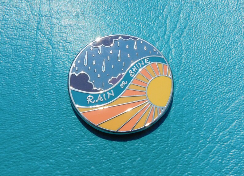 Puede incluir: Un pin redondo de color plateado con un cielo azul y nubes de lluvia en una mitad y un sol amarillo con rayos naranjas en la otra mitad. El texto "RAIN OR SHINE" est&aacute; escrito en una fuente blanca cursiva.