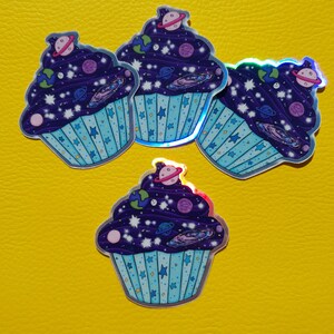 May include: Four holographic stickers of galaxy cupcakes. Each cupcake is decorated with stars, planets, and a swirl of galaxy colors. The cupcakes are on a yellow background.
