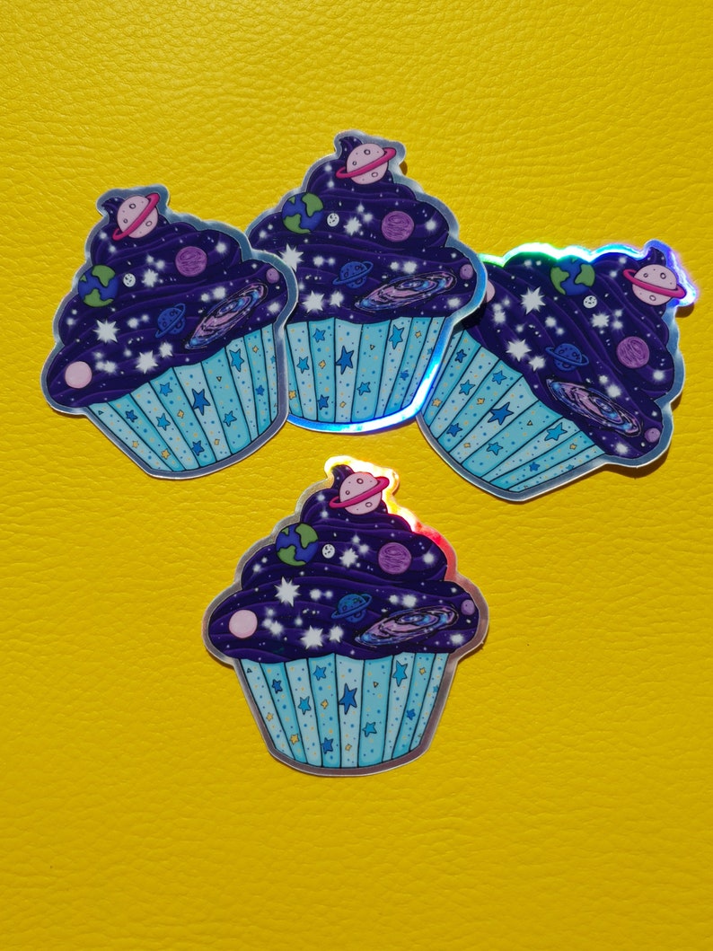 May include: Four holographic stickers of galaxy cupcakes. Each cupcake is decorated with stars, planets, and a swirl of galaxy colors. The cupcakes are on a yellow background.