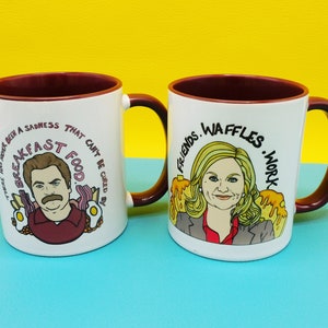 Puede incluir: Dos tazas de cerámica blanca con bordes rojos. Una taza presenta una ilustración de dibujos animados de Ron Swanson de Parks and Recreation con el texto "Nunca ha habido una tristeza que no pueda ser curada por la comida del desayuno." La otra taza presenta una ilustración de dibujos animados de Leslie Knope de Parks and Recreation con el texto "Friends. Waffles. Work."