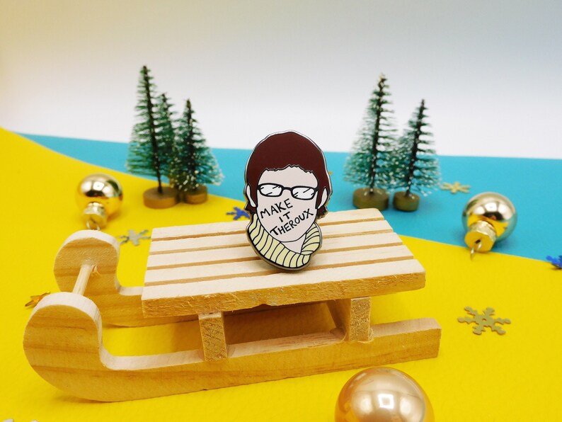 May include: A brown and white enamel pin featuring a cartoon illustration of a person with brown hair wearing glasses and a yellow scarf. The text "Make it Theroux" is written across the person's mouth.