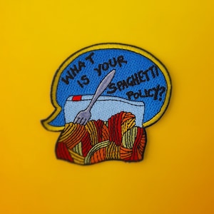 Puede incluir: Un parche bordado azul y amarillo con el texto "¿Cuál es tu política de espaguetis?" y un tenedor clavado en una caja de espaguetis.