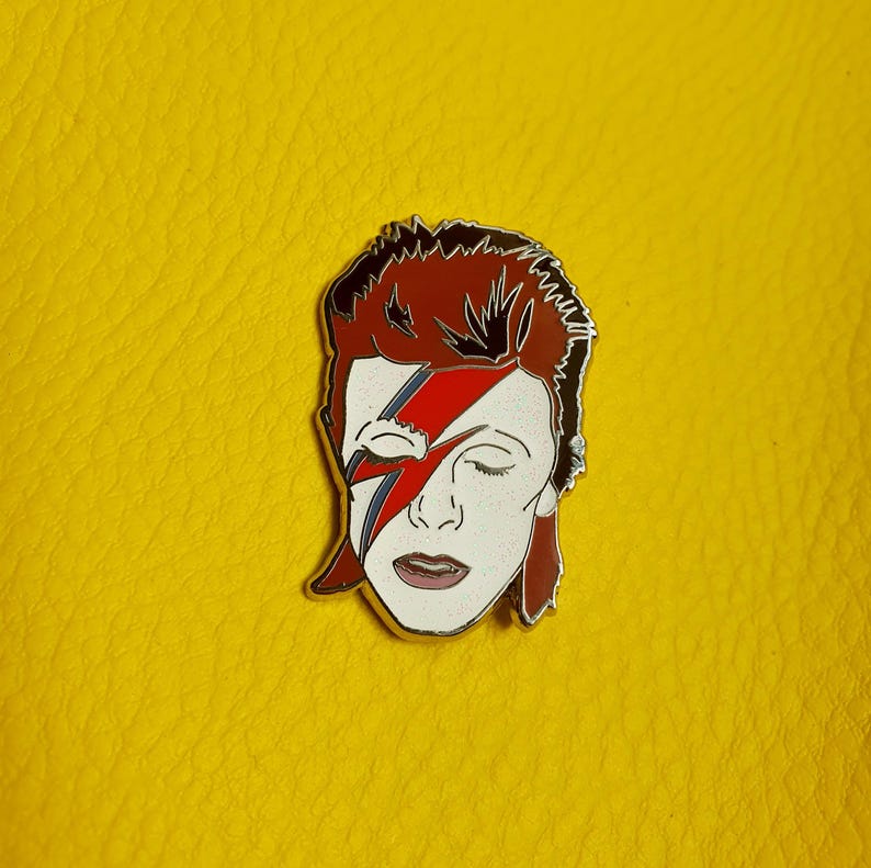 Puede incluir: Un pin de esmalte plateado con una ilustraci&oacute;n roja y negra de la cara de David Bowie con su caracter&iacute;stico maquillaje de rayo.