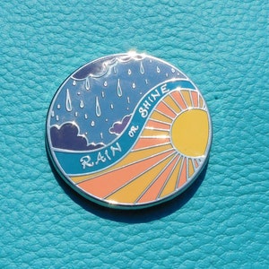Puede incluir: Un pin de esmalte redondo con un cielo azul y nubes de lluvia en un lado y un sol amarillo con rayos naranjas en el otro. El texto "RAIN or SHINE" est&aacute; escrito en una l&iacute;nea curva alrededor del sol.