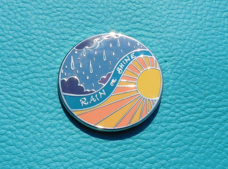 Puede incluir: Un pin de esmalte redondo con un cielo azul y nubes de lluvia en un lado y un sol amarillo con rayos naranjas en el otro. El texto "RAIN or SHINE" est&aacute; escrito en una l&iacute;nea curva alrededor del sol.