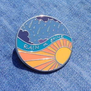 Puede incluir: Un pin de esmalte redondo con un cielo azul, nubes blancas y gotas de lluvia en la mitad superior. La mitad inferior es un sol amarillo con rayos naranjas. El texto "RAIN or SHINE" est&aacute; escrito en un arco blanco en el medio.