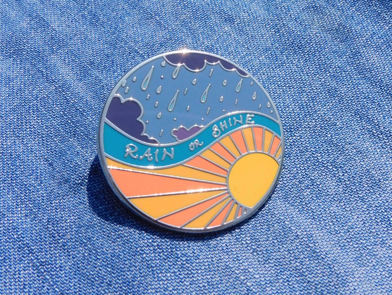 Puede incluir: Un pin de esmalte redondo con un cielo azul, nubes blancas y gotas de lluvia en la mitad superior. La mitad inferior es un sol amarillo con rayos naranjas. El texto "RAIN or SHINE" est&aacute; escrito en un arco blanco en el medio.