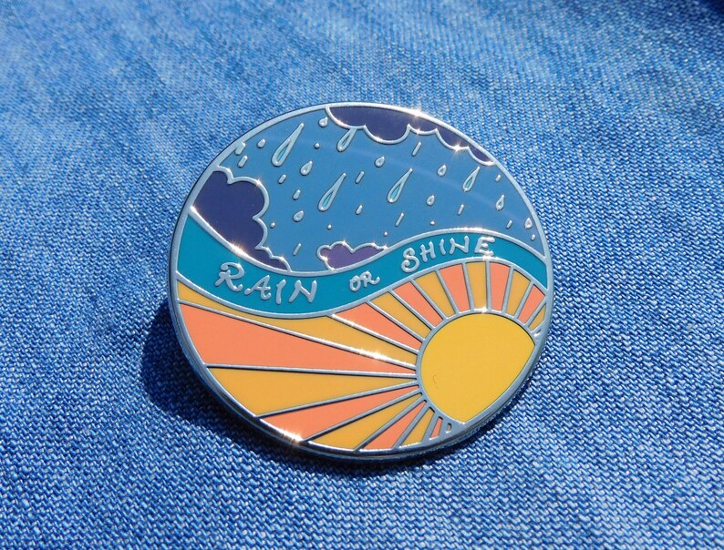 Puede incluir: Un pin de esmalte redondo con un cielo azul, nubes blancas y gotas de lluvia en la mitad superior. La mitad inferior es una salida del sol con colores naranja, amarillo y rosa. El texto "RAIN or SHINE" est&aacute; escrito en blanco sobre un fondo azul turquesa.