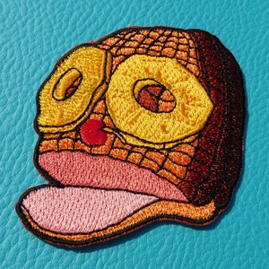 May include: Embroidered iron-on patch of a cartoon face with a large open mouth, yellow eyes, and a red nose. The face is brown, orange, and pink.