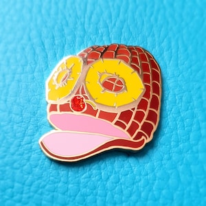 May include: A red and yellow enamel pin shaped like a pineapple with a pink tongue sticking out. The pineapple has a red cherry on top.
