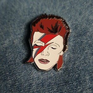 Puede incluir: Un pin de esmalte que representa un retrato de David Bowie con su ic&oacute;nico maquillaje de rayo, en rojo y blanco.