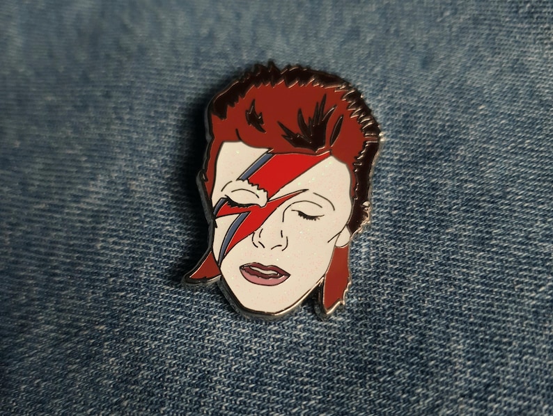 Puede incluir: Un pin de esmalte que representa un retrato de David Bowie con su ic&oacute;nico maquillaje de rayo, en rojo y blanco.